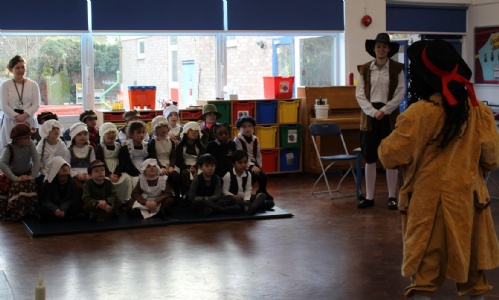 Children sitting in the school hall dressed in Victorian costume including white aprons, mop caps, flat caps and waistcoats.